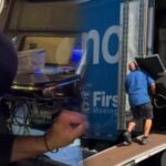 A repressão ao jogo na Flórida continua com a apreensão de outras 134 máquinas caça-níqueis. Duas cenas combinadas: à esquerda, uma pessoa com capuz parece inspecionar ou manipular uma máquina caça-níqueis em um ambiente escuro; à direita, um trabalhador de camisa azul carrega uma grande máquina de jogos em um caminhão em movimento usando uma rampa.