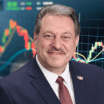 Addabbo abre revisão legal em Nova York para potencialmente regular os mercados de previsão. Um homem de terno azul-marinho e gravata vermelha sorri diante de um fundo que mostra gráficos financeiros coloridos e gráficos de velas.