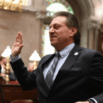 Addabbo exige uma regulamentação rápida em Nova Iorque à medida que os mercados de previsão se expandem rapidamente. O senador Addabbo de terno levanta a mão direita enquanto fala ao microfone dentro de uma câmara formal do governo com decoração ornamentada e vitrais.