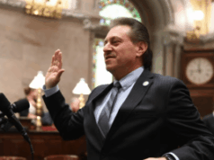 Addabbo exige regulamentação rápida de Nova York à medida que os mercados de previsão se expandem rapidamente Addabbo exige uma regulamentação rápida em Nova Iorque à medida que os mercados de previsão se expandem rapidamente. O senador Addabbo de terno levanta a mão direita enquanto fala ao microfone dentro de uma câmara formal do governo com decoração ornamentada e vitrais.