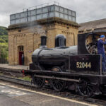 O interior do centro de visitantes. As paredes são pintadas de branco e construídas em pedra ou tijolo. O teto é arqueado e apresenta vigas expostas pintadas em cor escura, além de luminárias modernas penduradas. No lado esquerdo da imagem, há uma grande janela com vários painéis. Abaixo da janela, há vitrines contendo vários objetos, incluindo três estruturas altas e estreitas em vermelho, cinza e azul, que parecem ser equipamentos ferroviários históricos ou dispositivos de sinalização. Cada display possui etiquetas para identificação. No centro, em primeiro plano, há um elemento circular coberto de vidro inserido no chão. Perto da parede dos fundos, há uma porta vermelha brilhante que está aberta. Acima desta porta, há uma placa que diz “KEIGHLEY”. Abaixo da placa, três telas digitais são montadas na parede, mostrando imagens e textos relacionados à história ferroviária.