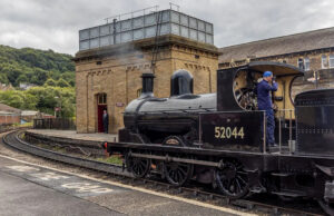 Centro de visitantes de torres de água raras será premiado O interior do centro de visitantes. As paredes são pintadas de branco e construídas em pedra ou tijolo. O teto é arqueado e apresenta vigas expostas pintadas em cor escura, além de luminárias modernas penduradas. No lado esquerdo da imagem, há uma grande janela com vários painéis. Abaixo da janela, há vitrines contendo vários objetos, incluindo três estruturas altas e estreitas em vermelho, cinza e azul, que parecem ser equipamentos ferroviários históricos ou dispositivos de sinalização. Cada display possui etiquetas para identificação. No centro, em primeiro plano, há um elemento circular coberto de vidro inserido no chão. Perto da parede dos fundos, há uma porta vermelha brilhante que está aberta. Acima desta porta, há uma placa que diz “KEIGHLEY”. Abaixo da placa, três telas digitais são montadas na parede, mostrando imagens e textos relacionados à história ferroviária.