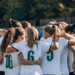Como a ascensão do futebol feminino está impactando as apostas esportivas. Um time de futebol feminino está reunido no campo, abraçados, vestindo camisas brancas com números verdes. Eles parecem concentrados e unidos, preparando-se juntos antes de uma partida, com um estádio desfocado e árvores ao fundo.