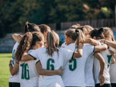 Como a ascensão do futebol feminino está impactando as apostas esportivas? Como a ascensão do futebol feminino está impactando as apostas esportivas. Um time de futebol feminino está reunido no campo, abraçados, vestindo camisas brancas com números verdes. Eles parecem concentrados e unidos, preparando-se juntos antes de uma partida, com um estádio desfocado e árvores ao fundo.