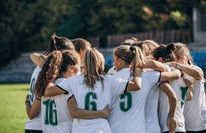 Como a ascensão do futebol feminino está impactando as apostas esportivas? Como a ascensão do futebol feminino está impactando as apostas esportivas. Um time de futebol feminino está reunido no campo, abraçados, vestindo camisas brancas com números verdes. Eles parecem concentrados e unidos, preparando-se juntos antes de uma partida, com um estádio desfocado e árvores ao fundo.