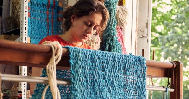 Nabil olha diretamente para a câmera, com um cachimbo na boca e na mão esquerda. Atrás dele está uma grande obra de arte em tons terrosos