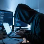 Uma fotografia sombria de uma figura sombria debruçada sobre um laptop brilhante em uma sala mal iluminada, com o rosto parcialmente obscurecido pela luz azul que emana da tela. Connecticut alerta sobre esquema DraftKings que leva a prisões em fraude crescente