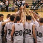 Time de basquete Coopersville Broncos. Um time de basquete de uma escola ou faculdade se reúne na quadra central com as mãos levantadas antes do jogo. Os jogadores vestem camisetas brancas combinando com detalhes escuros, e os espectadores sentam-se nas arquibancadas ao fundo. Descobriu-se que seis estudantes atletas manipularam apostas pela NCAA