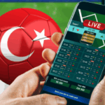 Uma pessoa segurando um smartphone exibindo uma tela de apostas esportivas ao vivo, com uma bola de futebol vermelha com a bandeira turca ao fundo, sugerindo atividades de apostas relacionadas à liga turca de futebol. Escândalo com árbitros da liga turca faz com que mais de cem dirigentes sejam suspensos