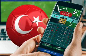 Escândalo com árbitros da liga turca faz com que mais de cem dirigentes sejam suspensos Uma pessoa segurando um smartphone exibindo uma tela de apostas esportivas ao vivo, com uma bola de futebol vermelha com a bandeira turca ao fundo, sugerindo atividades de apostas relacionadas à liga turca de futebol. Escândalo com árbitros da liga turca faz com que mais de cem dirigentes sejam suspensos