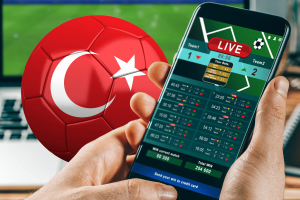 Escândalo com árbitros da liga turca faz com que mais Uma pessoa segurando um smartphone exibindo uma tela de apostas esportivas ao vivo, com uma bola de futebol vermelha com a bandeira turca ao fundo, sugerindo atividades de apostas relacionadas à liga turca de futebol. Escândalo com árbitros da liga turca faz com que mais de cem dirigentes sejam suspensos