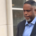 O ex-astro da NBA Damon Jones se declara inocente em casos de conspiração de jogos de azar e pôquer. Um homem de terno escuro e camisa azul clara sai de um prédio, olhando para baixo com uma expressão séria.