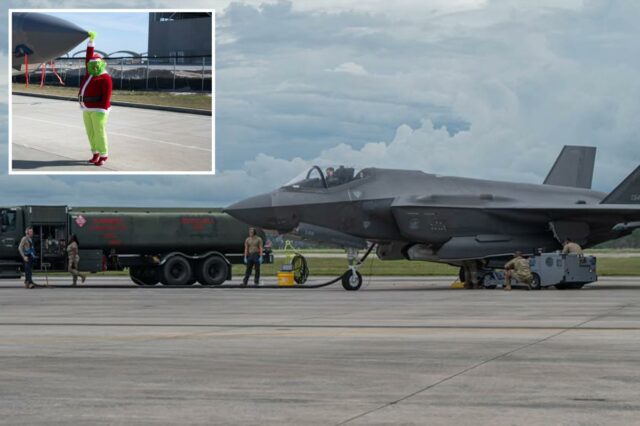 Uma pessoa fantasiada de Grinch com chapéu e terno de Papai Noel segura o nariz de um jato militar cinza estacionado.