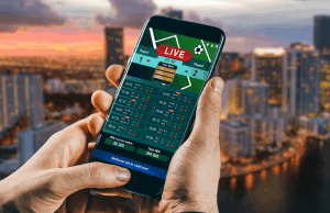 Florida House avalia revisão dos jogos de azar, endurecendo as penalidades e regulamentando os esportes de fantasia Florida House avalia revisão dos jogos de azar, endurecendo as penalidades e regulamentando os esportes de fantasia. Uma pessoa segurando um smartphone exibindo uma interface de apostas esportivas ao vivo, com o horizonte de uma cidade desfocado ao fundo ao pôr do sol.