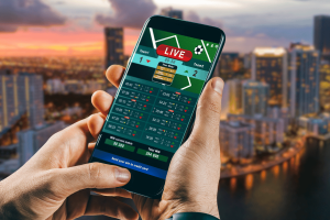 Florida House avalia revisão dos jogos de azar, endurecendo as Florida House avalia revisão dos jogos de azar, endurecendo as penalidades e regulamentando os esportes de fantasia. Uma pessoa segurando um smartphone exibindo uma interface de apostas esportivas ao vivo, com o horizonte de uma cidade desfocado ao fundo ao pôr do sol.