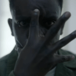 Um homem olha para a câmera com a mão sobre o rosto e os dedos afastados.