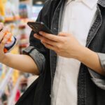 Estudante chinês asiático olhando para smartphone enquanto escolhe itens no supermercado.