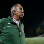 John Beam, com uma jaqueta verde, olhando para um campo de futebol à noite.
