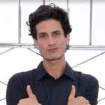 Jack Schlossberg está diante de várias barras de metal. Seus braços estão cruzados sobre o peito e ele está esticando o polegar e o dedo mínimo.