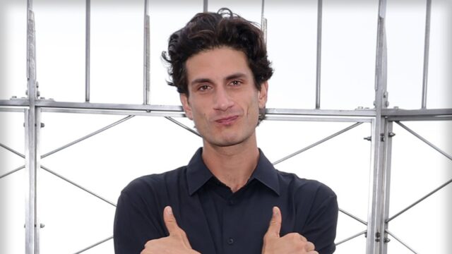 Jack Schlossberg está diante de várias barras de metal. Seus braços estão cruzados sobre o peito e ele está esticando o polegar e o dedo mínimo.
