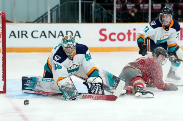 A goleira do New York Sirens, Kayle Osborne (82), observa o disco enquanto Brianne Jenner (19), do Ottawa Charge, desliza ao longo do gelo.