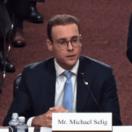 O indicado à CFTC, Michael Selig, evita as críticas dos senadores, mantendo os mercados de previsões esportivas no limbo legal. Michael Selig está sentado em uma mesa de audiência no Senado, vestindo terno escuro, gravata azul clara e óculos, falando em um microfone com uma placa onde se lê “Sr. Michael Selig” à sua frente.