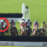 O painel judicial do BHA atinge Kitts e Higgins com grandes proibições no escândalo de apostas em Hillsin. Um grupo de jóqueis a cavalo pula uma cerca durante uma corrida em uma pista gramada. Um cavalo e um cavaleiro em sedas brancas e vermelhas são destacados com um círculo vermelho em primeiro plano.