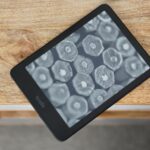 Kindle Paperwhite (2024) deitado na borda da mesa mostrando uma tela com seleção de livros