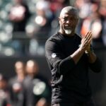 Um jogador do Cleveland Browns com uniforme branco segurando uma bola de futebol foge de um jogador do New England Patriots com uniforme azul.
