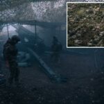 Dois soldados, vestidos com camuflagem, estão sob uma rede de camuflagem ao lado de um canhão de artilharia.