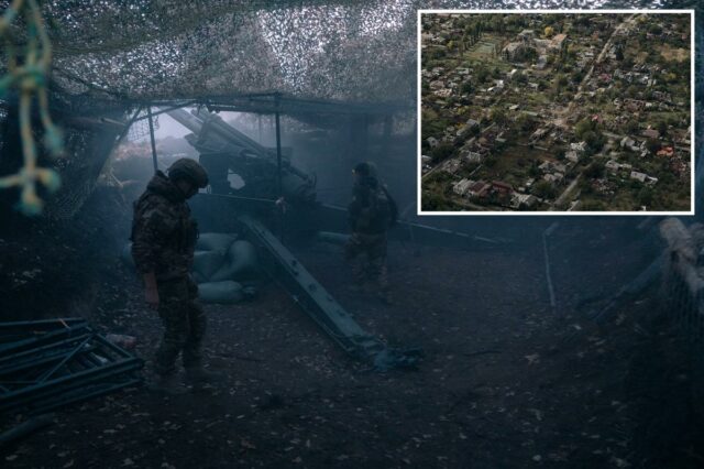 Dois soldados, vestidos com camuflagem, estão sob uma rede de camuflagem ao lado de um canhão de artilharia.