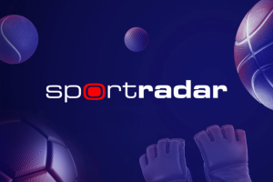 Sportradar adquire IMG ARENA para impulsionar esforços globais de expansão Logotipo da Sportradar exibido em um fundo azul escuro com elementos esportivos estilizados, incluindo bola de tênis, bola de golfe, basquete, bola de futebol e luvas de goleiro. Sportradar adquire IMG ARENA para impulsionar esforços globais de expansão de dados esportivos