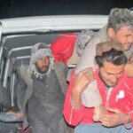 Uma mulher idosa está sentada em uma maca em um hospital. Uma bandagem cobre a cabeça e os olhos. Uma mulher segurando uma criança observa.