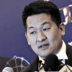 O tribunal tailandês ordena que o chefão do jogo seja extraditado para a China em meio a uma longa batalha legal. She Zhijiang de terno fala em vários microfones durante um evento de imprensa, em frente a um fundo azul com texto e gráficos.