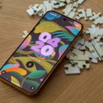 Mascote do Android entre caixas de papelão etiquetadas para aplicativos, capturas de tela, notificações e distrações, em frente à tela de um smartphone, com uma vassoura apoiada nas caixas.