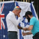 Uma bandeira israelense colocada no chão entre flores, homenagens e um pequeno caderno com canetas.