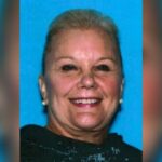 Mugshot de uma mulher sorridente com cabelos claros em um top preto contra um fundo azul.