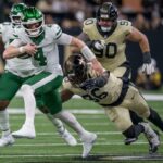 O quarterback do New York Jets, Brady Cook (4), corre com a bola enquanto o lado defensivo do New Orleans Saints, Carl Granderson (96), enfrenta outro jogador do Saints no primeiro tempo de um jogo de futebol.