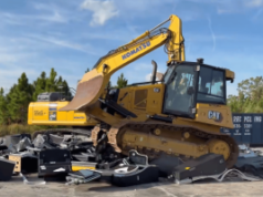 Comissão de Controle de Jogos da Flórida destrói 147 caça-níqueis ilegais A Comissão de Controle de Jogos da Flórida destrói 147 caça-níqueis ilegais. Uma escavadeira CAT amarela passa e esmaga uma grande pilha de eletrônicos quebrados e detritos de metal em um terreno de concreto. Atrás dela, uma escavadeira Komatsu amarela está estacionada perto de uma área arborizada sob um céu azul.