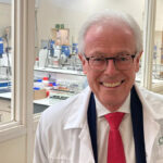 Um casal de meia-idade está em frente a um conjunto de portas brancas segurando uma claquete de filme e um recorte de revista. À esquerda, um homem com cabelos escuros e grisalhos, vestindo um blazer fulvo e com o polegar para cima. À direita uma mulher de cabelos castanhos, amarrados para trás, vestindo um suéter azul e rosa. A claquete é branca com as palavras "Óleo de Lorenzo, diretor George Miller, câmera John Seale" impresso nele em letras vermelhas. O recorte da revista inclui a foto de um menino vestindo uma camisa de rugby listrada de vermelho e branco.