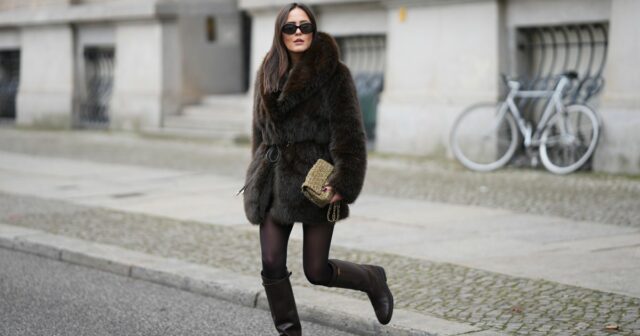 Um convidado usa óculos de sol circulares, um cardigã curto vermelho fofo de mangas curtas / abotoado, calça jeans azul marinho de pernas largas, uma bolsa cesta de vime marrom, um lenço de seda com estampa verde / azul / laranja, anéis, sapatos de salto de couro envernizado branco brilhante, um cinto de corrente de ouro, fora do desfile da Hermes, durante a Paris Fashion Week - Menswear Primavera/Verão 2023, em 25 de junho de 2022 em Paris, França.