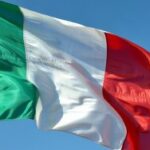 Uma fotografia em close da bandeira italiana ondulando dramaticamente sob uma forte brisa contra um céu azul claro. O tecido tricolor apresenta listras verticais em verde esmeralda vívido, branco imaculado e vermelho carmesim profundo, ondulando com movimento dinâmico, criando ondas e dobras naturais que captam a luz. A textura da bandeira é bem visível, mostrando a trama do tecido à medida que se estica e se contrai com o vento. A forte luz do sol mediterrâneo ilumina as cores por trás, fazendo-as parecer luminosas e saturadas, ao mesmo tempo que lança sombras sutis nos vincos do tecido.