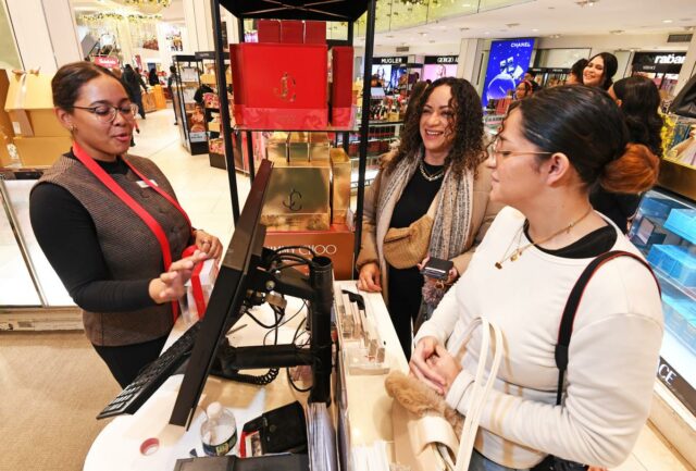 Funcionária da Macy's ajudando duas mulheres nas compras de Natal de última hora em uma loja de perfumes.
