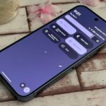 Um grande ícone do Quick Share centralizado em um fundo verde claro, com caminhos curvos pontilhados conectando mascotes do Android, pastas de documentos e ícones de dispositivos como telefone, tablet e laptop.