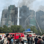 O Jockey Club de Hong Kong realiza corridas a portas fechadas e doa os lucros às vítimas dos incêndios. Os bombeiros borrifam água em vários arranha-céus residenciais fortemente danificados e enegrecidos por um grande incêndio, com fumaça subindo dos edifícios. Veículos de emergência e caminhões de bombeiros estão reunidos abaixo, enquanto equipes de mídia e curiosos ficam atrás de barreiras observando a cena.