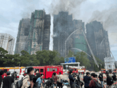 O Jockey Club de Hong Kong realiza corrida a portas fechadas e doa lucros às vítimas do incêndio O Jockey Club de Hong Kong realiza corridas a portas fechadas e doa os lucros às vítimas dos incêndios. Os bombeiros borrifam água em vários arranha-céus residenciais fortemente danificados e enegrecidos por um grande incêndio, com fumaça subindo dos edifícios. Veículos de emergência e caminhões de bombeiros estão reunidos abaixo, enquanto equipes de mídia e curiosos ficam atrás de barreiras observando a cena.