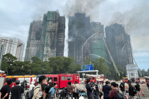 O Jockey Club de Hong Kong realiza corrida a portas O Jockey Club de Hong Kong realiza corridas a portas fechadas e doa os lucros às vítimas dos incêndios. Os bombeiros borrifam água em vários arranha-céus residenciais fortemente danificados e enegrecidos por um grande incêndio, com fumaça subindo dos edifícios. Veículos de emergência e caminhões de bombeiros estão reunidos abaixo, enquanto equipes de mídia e curiosos ficam atrás de barreiras observando a cena.