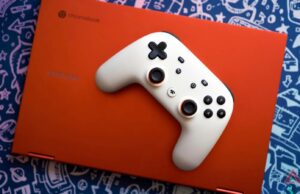 O controlador Stadia do Google acaba de receber uma grande atualização, cortesia do Steam Um controlador branco do Google Stadia no teclado de um Chromebook vermelho com um serviço de jogos aberto na tela. O laptop está sobre um fundo azul com gráficos prateados de imagens de videogame.