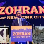 Zohran Mamdani comemora depois de garantir sua vitória para prefeito da cidade de Nova York, falando em um pódio com “Zohran pela cidade de Nova York” e uma bandeira americana à sua esquerda.