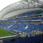 Estádio AMEX de futebol Brighton & Hove Albion, campo verde à esquerda e cadeiras à direita. O presidente do futebol, Tony Bloom, foi alvo de acusações de apostas, mas nega as acusações